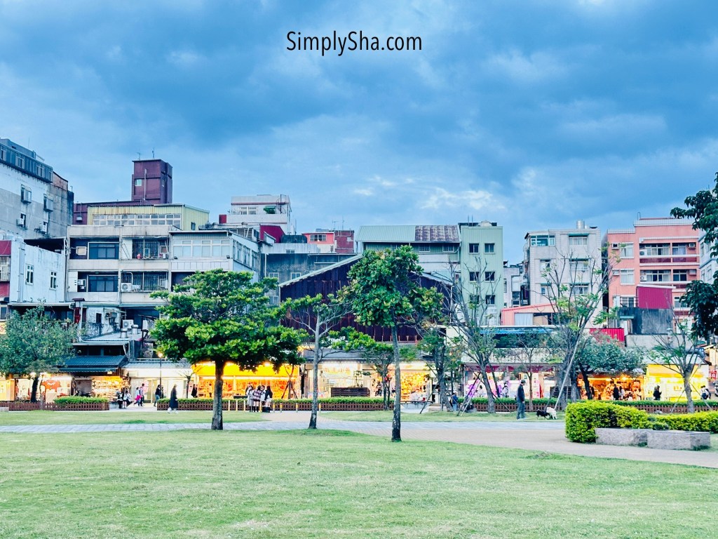 Open park area near Tamsui Old Street with grassy space and surrounding buildings at dusk