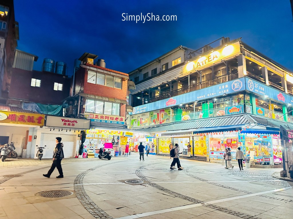 Tamsui Old Street at night with brightly lit shops, street food stalls, and pedestrians